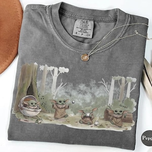 May include: A gray Comfort Colors t-shirt featuring a Grogu graphic in a forest scene. The design includes Grogu in a pod, and Grogu drinking tea. A silver necklace with a pendant is on the shirt. The shirt has a small tag.