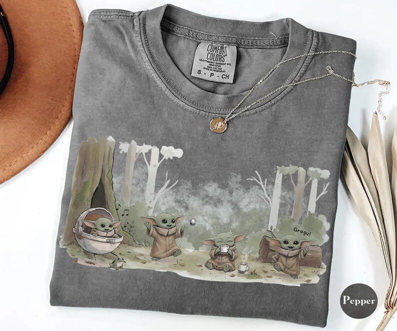 May include: A gray Comfort Colors t-shirt featuring a Grogu graphic in a forest scene. The design includes Grogu in a pod, and Grogu drinking tea. A silver necklace with a pendant is on the shirt. The shirt has a small tag.