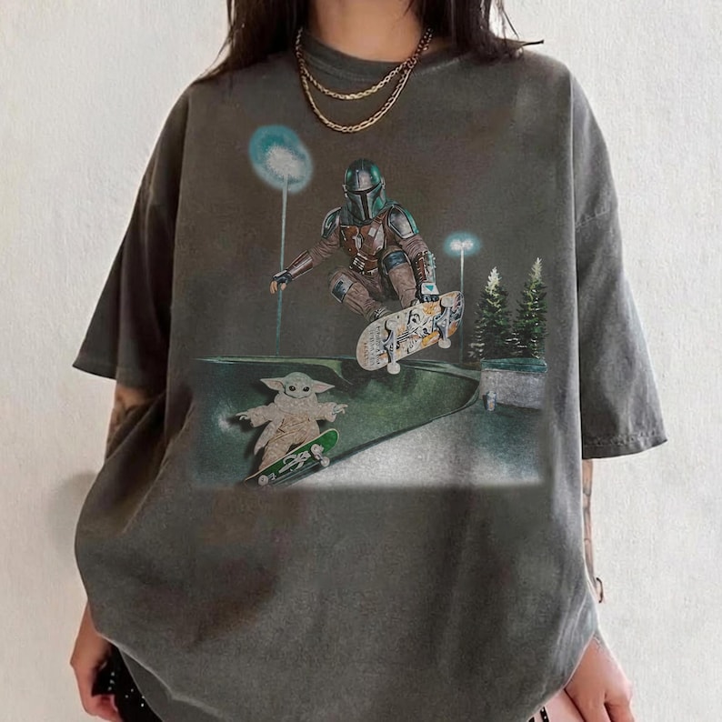 May include: Dark gray t-shirt featuring a graphic of The Mandalorian and Grogu skateboarding in a skate park. The design includes a night scene with streetlights and trees. The shirt has a vintage, worn look.