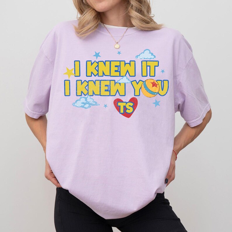 May include: A light purple short-sleeve t-shirt featuring the yellow text I KNEW IT I KNEW YOU with a red heart, a yellow star, blue clouds, and a yellow ball with a red star. The design includes the letters TS inside the red heart graphic.