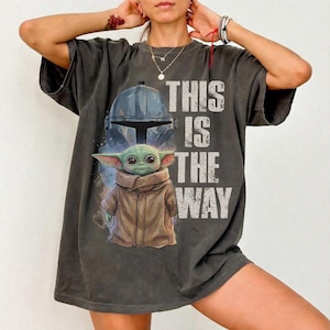 Peut inclure: T-shirt gris fonc&eacute; avec un motif d'un casque Mandalorien et de Grogu, avec le texte "THIS IS THE WAY" en blanc. Le design comprend un casque bleu clair et un Grogu vert portant un manteau marron.