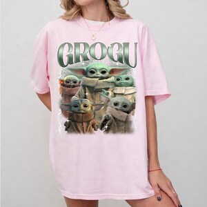 K&ouml;nnte beinhalten: Ein hellrosa T-Shirt mit einer Grafik von Grogu, auch bekannt als Baby Yoda, aus The Mandalorian. Die Grafik enth&auml;lt das Wort "GROGU" in einer gr&uuml;nen, metallischen Schrift &uuml;ber mehreren Bildern von Grogu.