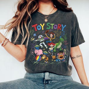 Puede incluir: Camiseta gris oscuro con un gr&aacute;fico colorido de los personajes de Toy Story. El dise&ntilde;o incluye a Buzz Lightyear, Woody y otros personajes de la pel&iacute;cula animada, con las palabras "TOY STORY" en una fuente l&uacute;dica.