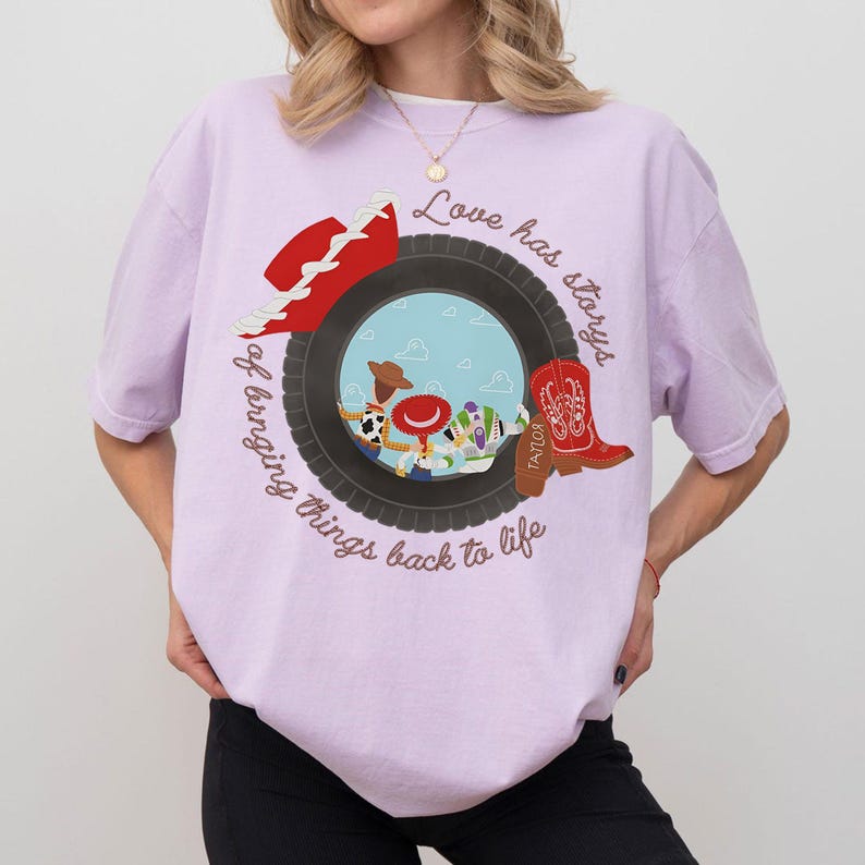 May include: A light purple t-shirt featuring a graphic of a black tire framing a scene with three cartoon characters. The design includes a red cowboy hat, a red cowboy boot with the name Taylor, and the text Love has storys of bringing things back to life.