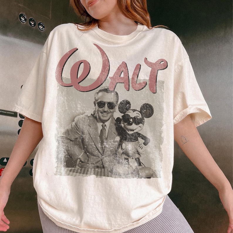May include: Cream-colored t-shirt featuring a vintage-style graphic of Walt Disney and Mickey Mouse. The word "Walt" is printed in a pink, cursive font above the image. The image is a black and white photograph of Walt Disney and Mickey Mouse.