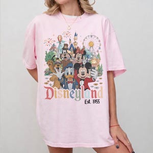 May include: A light pink t-shirt featuring a colorful graphic print of cartoon characters including Mickey Mouse, Minnie Mouse, Donald Duck, Goofy, and Pluto in front of a castle and ferris wheel. The text Disneyland Est. 1955 is printed below.