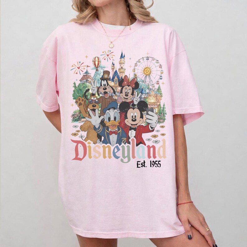 May include: A light pink t-shirt featuring a colorful graphic print of cartoon characters including Mickey Mouse, Minnie Mouse, Donald Duck, Goofy, and Pluto in front of a castle and ferris wheel. The text Disneyland Est. 1955 is printed below.