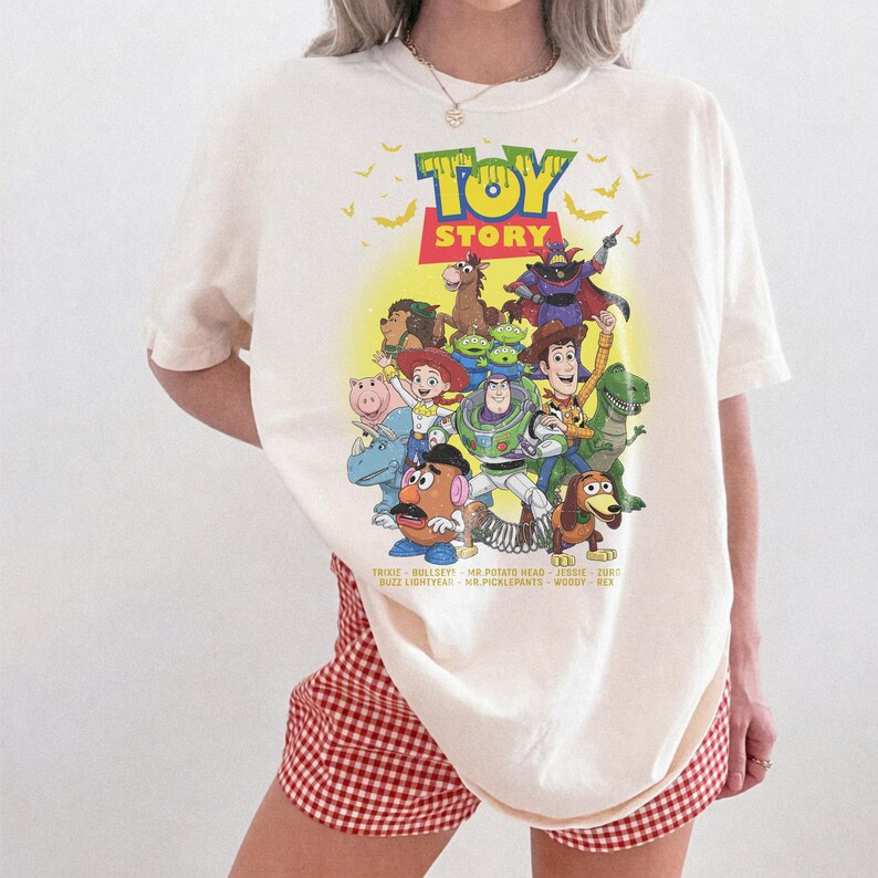 May include: An off-white short-sleeve t-shirt featuring a colourful Toy Story graphic print. The design includes characters like Woody, Buzz Lightyear, Jessie, and Zurg, with the Toy Story logo and small bats above, plus character names listed below.