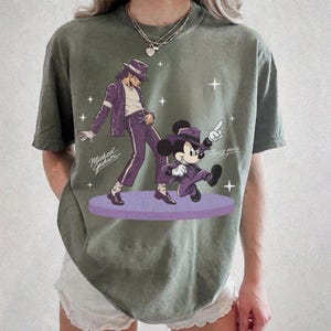 May include: Olive green t-shirt featuring a graphic of Michael Jackson and Mickey Mouse in purple outfits. The text "Michael Jackson" and "Mickey Mouse" are also visible. The design is on a purple stage with star accents.
