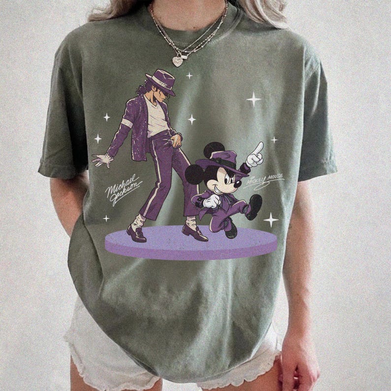 May include: Olive green t-shirt featuring a graphic of Michael Jackson and Mickey Mouse in purple outfits. The text "Michael Jackson" and "Mickey Mouse" are also visible. The design is on a purple stage with star accents.