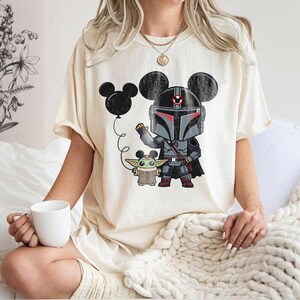 May include: Cream-colored t-shirt featuring a cartoon illustration of a Mandalorian character and Grogu, both with Mickey Mouse ears. A black balloon is also in the image. The shirt is a relaxed fit.
