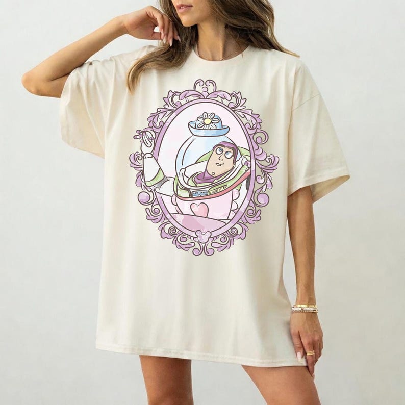 May include: Cream-colored t-shirt featuring a cartoon image of Buzz Lightyear inside a decorative oval frame. The frame is outlined in purple and pink, with floral and swirl details. The graphic is centered on the front of the shirt.