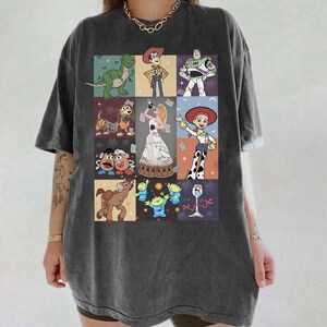 May include: A charcoal gray short-sleeve t-shirt featuring a grid of Toy Story characters including Woody, Buzz Lightyear, Rex, Slinky Dog, Mr. and Mrs. Potato Head, Bullseye, the Aliens, Forky, Jessie, and a central figure in a white gown.