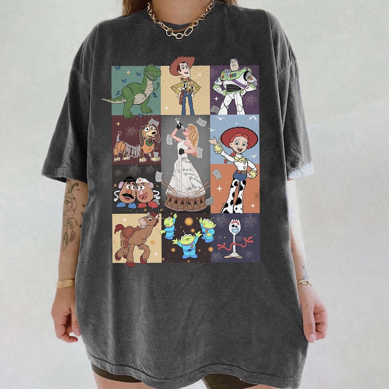 May include: A charcoal gray short-sleeve t-shirt featuring a grid of Toy Story characters including Woody, Buzz Lightyear, Rex, Slinky Dog, Mr. and Mrs. Potato Head, Bullseye, the Aliens, Forky, Jessie, and a central figure in a white gown.