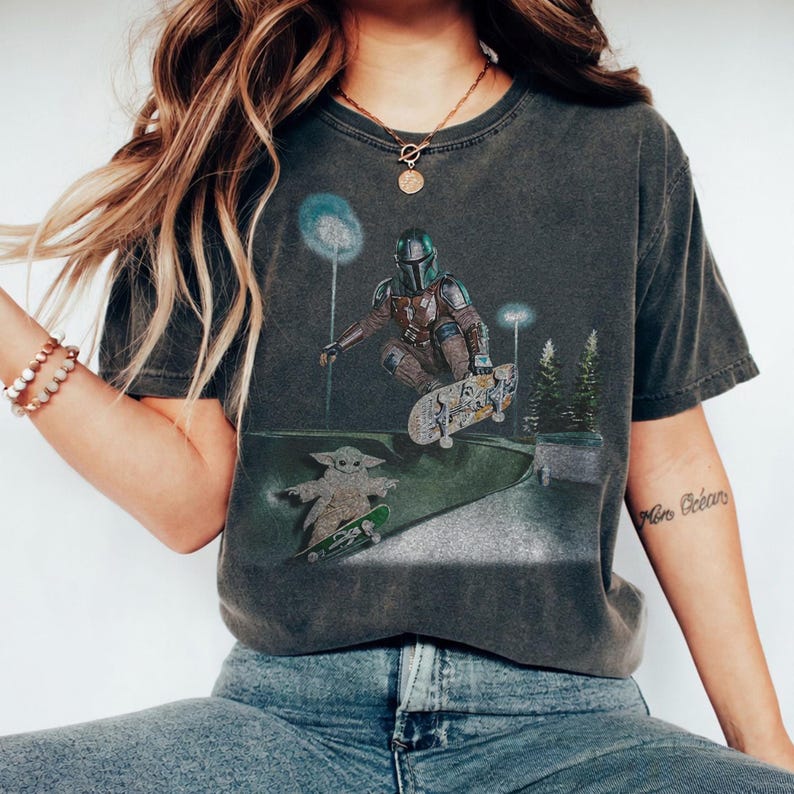 May include: Dark gray t-shirt featuring a graphic of a character from a popular science fiction series skateboarding. The design includes a character in armor and a small green creature on skateboards in a skate park setting.