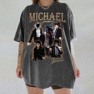 May include: Dark gray t-shirt featuring multiple images of Michael Jackson with the text "MICHAEL Jackson" in gold lettering. The shirt has a vintage, distressed look.