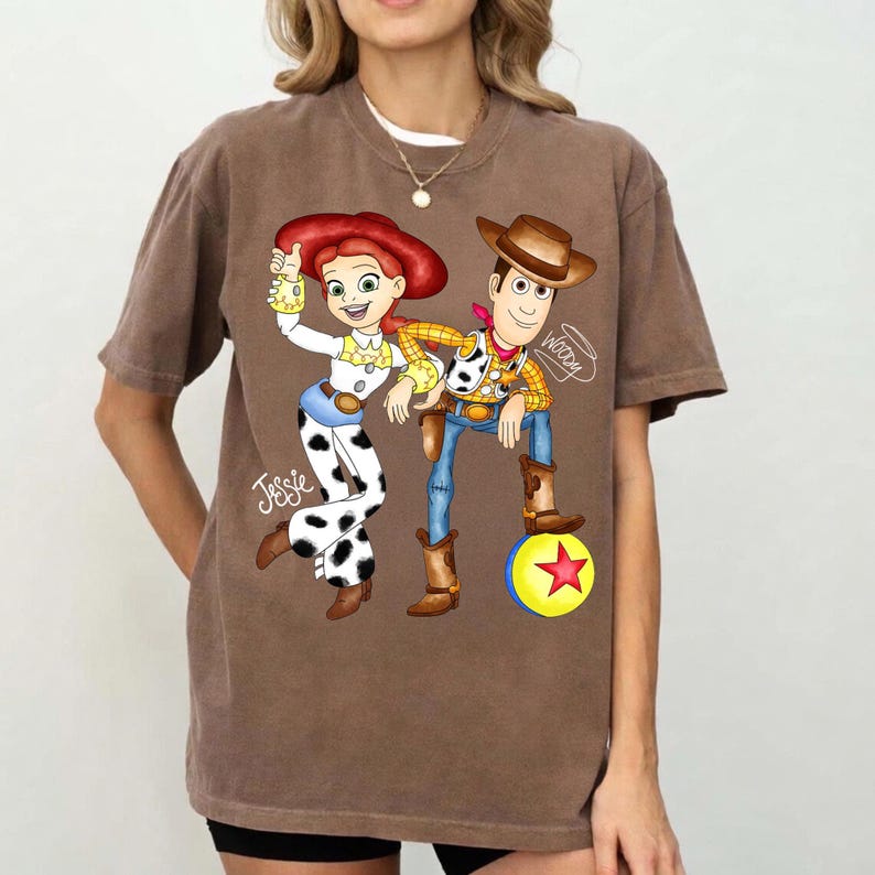 May include: Brown t-shirt with a cartoon illustration of Jessie and Woody from Toy Story. Jessie is wearing a white outfit with black cow-print trousers. Woody is wearing a cowboy hat and boots. The word "Woody" is written on the shirt.