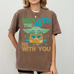 K&ouml;nnte beinhalten: Braunes T-Shirt mit einer gr&uuml;nen Alien-Grafik und dem Text "MAY THE 4TH BE WITH YOU" in Gr&uuml;n und Blau. Das Design enth&auml;lt Sterne und Kreuzformen.