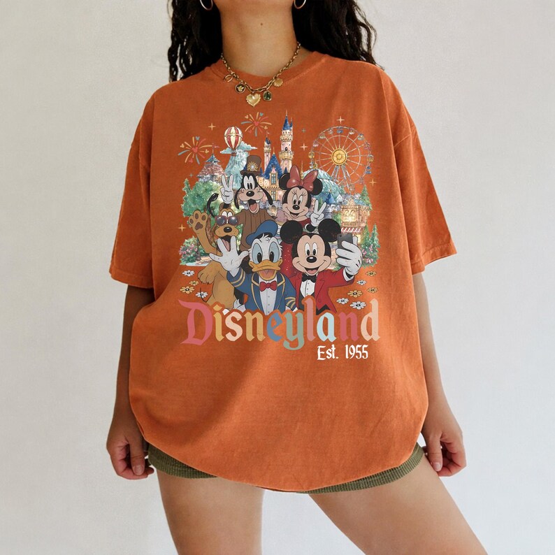 May include: An orange short-sleeve t-shirt featuring a graphic print of Mickey Mouse, Minnie Mouse, Donald Duck, Goofy, and Pluto at Disneyland. The text reads Disneyland Est. 1955, with a background illustration of a castle and a ferris wheel.
