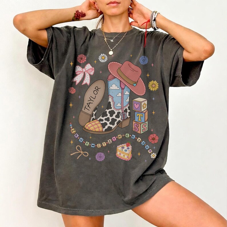 May include: A charcoal gray t-shirt featuring a graphic print of a cowboy boot with a cow print pattern, a pink hat, alphabet blocks spelling TOSS, a slice of cake, a bow, and the name TAYLOR. The design includes small stars and flowers.