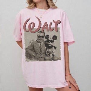 May include: Light pink t-shirt featuring a vintage-style graphic of Walt Disney with Mickey Mouse. The word "Walt" is printed in a cursive font above the image. The graphic is in shades of brown and gray.