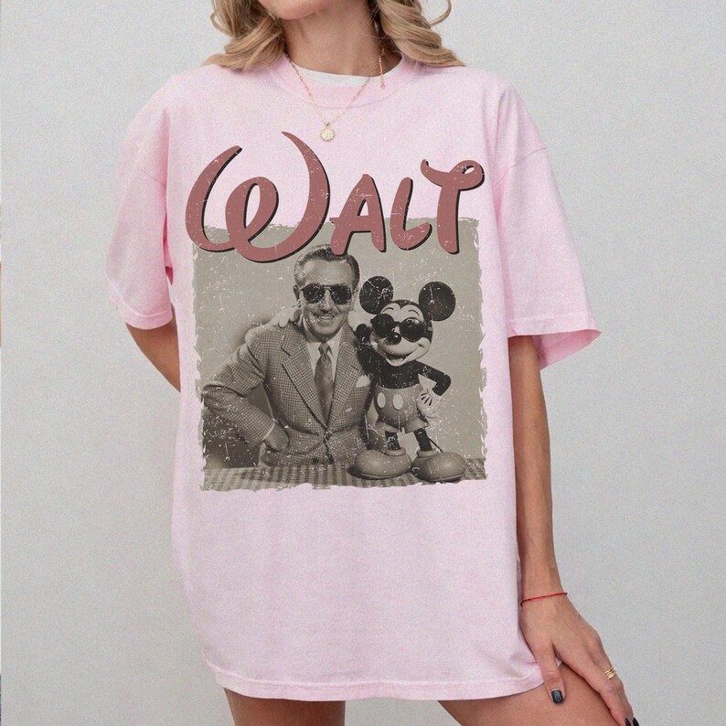 May include: Light pink t-shirt featuring a vintage-style graphic of Walt Disney with Mickey Mouse. The word "Walt" is printed in a cursive font above the image. The graphic is in shades of brown and gray.