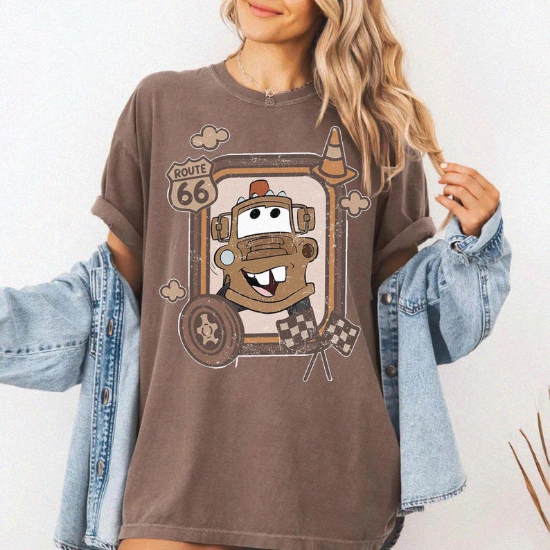 May include: Brown t-shirt featuring a cartoon image of Tow Mater from the movie Cars, framed by a Route 66 sign and racing flags. The design includes a tire, a traffic cone, and clouds. The shirt has rolled-up sleeves.