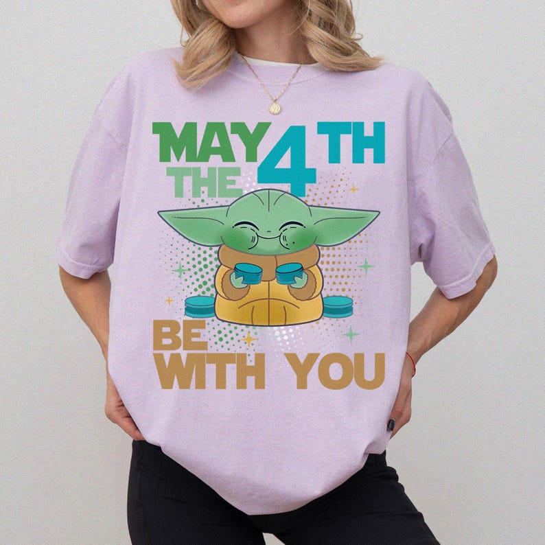 K&ouml;nnte beinhalten: Helllila T-Shirt mit einer Grafik eines gr&uuml;nen Alien-Charakters, der zwei blaue Tassen h&auml;lt. Der Text "MAY THE 4TH BE WITH YOU" ist oberhalb und unterhalb der Figur in Gr&uuml;n, Blau und Gold aufgedruckt.