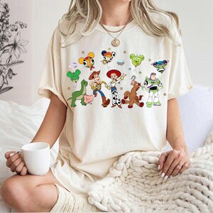 May include: Cream-colored t-shirt featuring a colorful graphic of Toy Story characters, including Woody, Buzz Lightyear, and others. The design includes balloons and stars. The shirt has short sleeves and a relaxed fit.