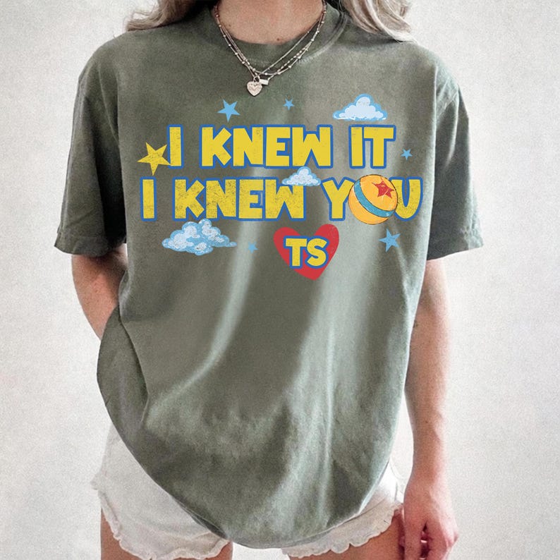 May include: An olive green short-sleeve t-shirt featuring the yellow text I KNEW IT I KNEW YOU, a red heart with the letters TS, a yellow ball with a red star, and light blue clouds and stars printed on the front of the garment.