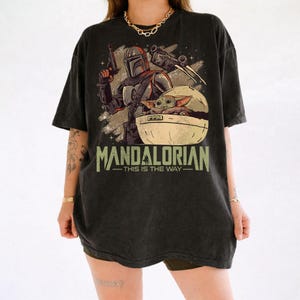May include: A black short-sleeve t-shirt featuring a graphic print of a Mandalorian character holding a blaster and a small creature in a floating pod. The text "MANDALORIAN" and "THIS IS THE WAY" is printed in a distressed green font below the illustration.