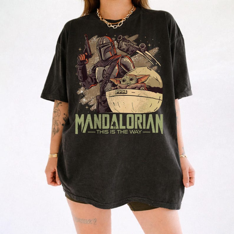 May include: A black short-sleeve t-shirt featuring a graphic print of a Mandalorian character holding a blaster and a small creature in a floating pod. The text "MANDALORIAN" and "THIS IS THE WAY" is printed in a distressed green font below the illustration.
