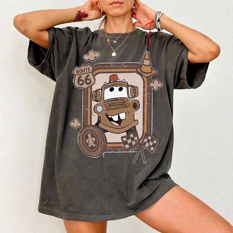 May include: Dark grey t-shirt featuring a cartoon image of Tow Mater from the movie Cars, framed by a Route 66 sign. The design includes a tire, clouds, and checkered flags. The shirt has short sleeves.