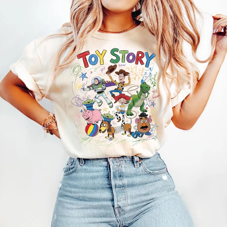Puede incluir: Camiseta color crema con las palabras "Toy Story" en letras coloridas y un gr&aacute;fico de dibujos animados de personajes de la pel&iacute;cula. El gr&aacute;fico incluye a Woody, Buzz Lightyear y otros personajes.