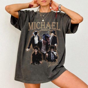 May include: Dark gray t-shirt featuring a graphic of Michael Jackson in various poses, with the name "MICHAEL Jackson" in gold lettering. The shirt has a vintage, worn look.