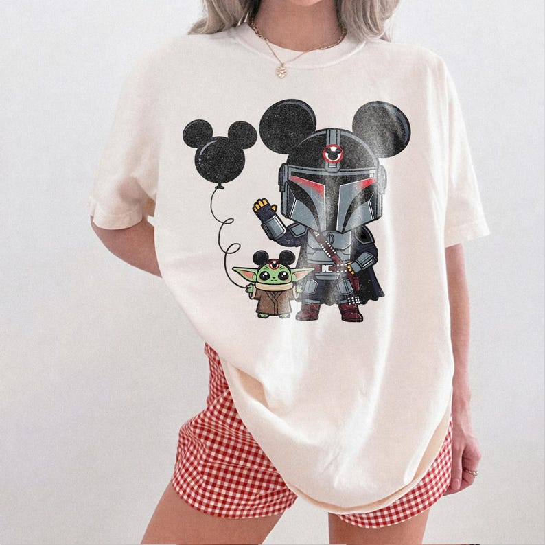 May include: Cream-colored t-shirt featuring a cartoon illustration of The Mandalorian and Grogu, with Mickey Mouse ears. The Mandalorian is in his armor, holding a black balloon. Grogu is standing next to him. The shirt has a relaxed fit.