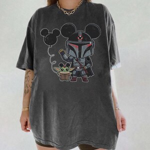 May include: Dark gray t-shirt featuring a cartoon illustration of a Mandalorian character with Mickey Mouse ears, holding a balloon with Mickey Mouse ears, and Grogu. The design is in black, gray, red, and brown.