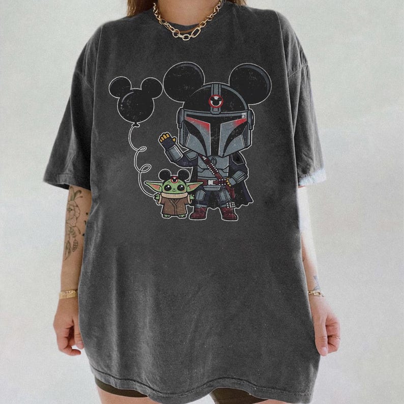 May include: Dark gray t-shirt featuring a cartoon illustration of a Mandalorian character with Mickey Mouse ears, holding a balloon with Mickey Mouse ears, and Grogu. The design is in black, gray, red, and brown.