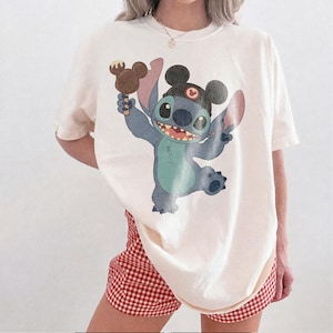 May include: Cream-colored t-shirt featuring a cartoon Stitch character holding a Mickey Mouse ice cream bar. Stitch is blue with large ears and a mischievous grin. The shirt is paired with red and white checkered shorts.
