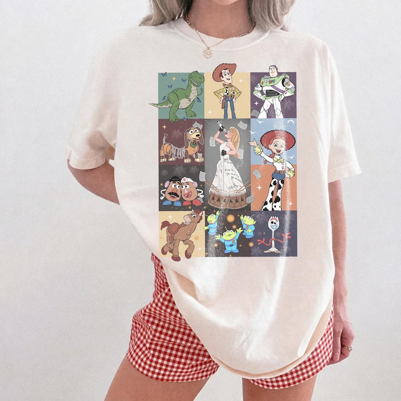 May include: A cream-colored short-sleeve t-shirt featuring a grid graphic print of various animated characters, including a cowboy, a space ranger, a cowgirl, a dinosaur, a dog, a potato couple, a horse, three aliens, and a white plastic utensil character.
