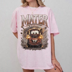 Puede incluir: Camiseta rosa claro con un dise&ntilde;o de Mate de la pel&iacute;cula Cars. La palabra "MATER" est&aacute; impresa encima del personaje en una fuente desgastada. El dise&ntilde;o incluye rayos y herramientas.