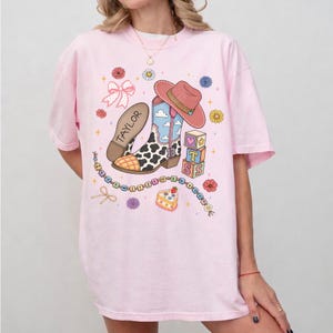 May include: A light pink t-shirt featuring a graphic print of a cowboy boot with a cow print pattern, a pink cowboy hat, a bow, alphabet blocks spelling TS, a cake, and a beaded string with the text I KNEW IT I KNEW IT. The boot has the name TAYLOR.