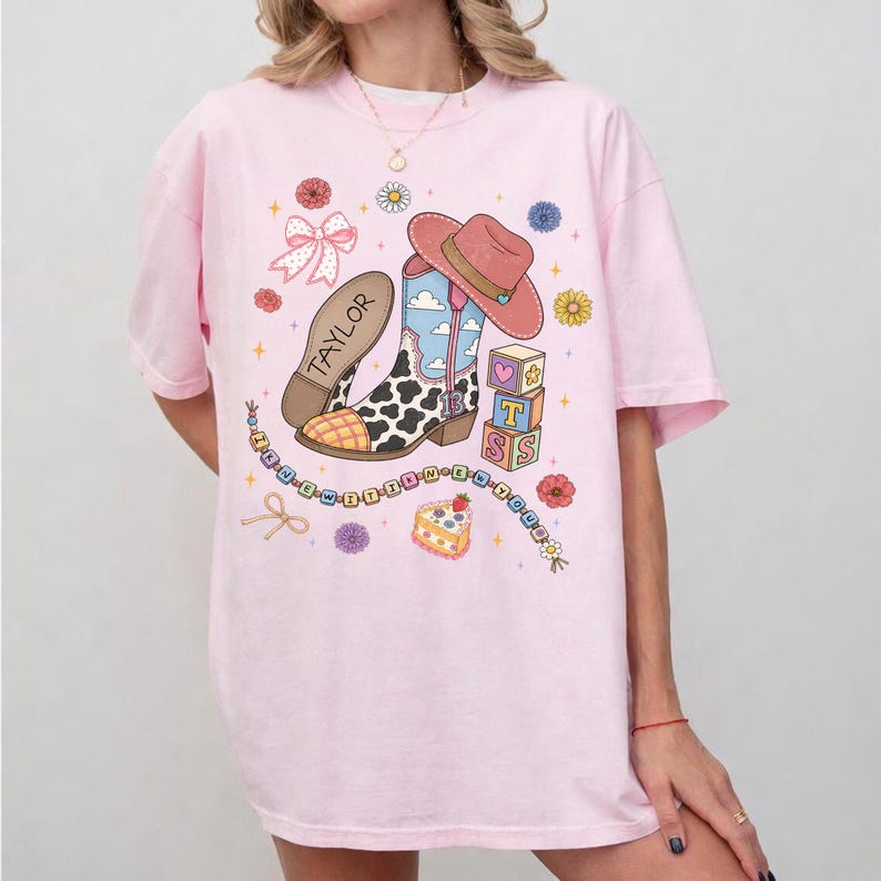 May include: A light pink t-shirt featuring a graphic print of a cowboy boot with a cow print pattern, a pink cowboy hat, a bow, alphabet blocks spelling TS, a cake, and a beaded string with the text I KNEW IT I KNEW IT. The boot has the name TAYLOR.