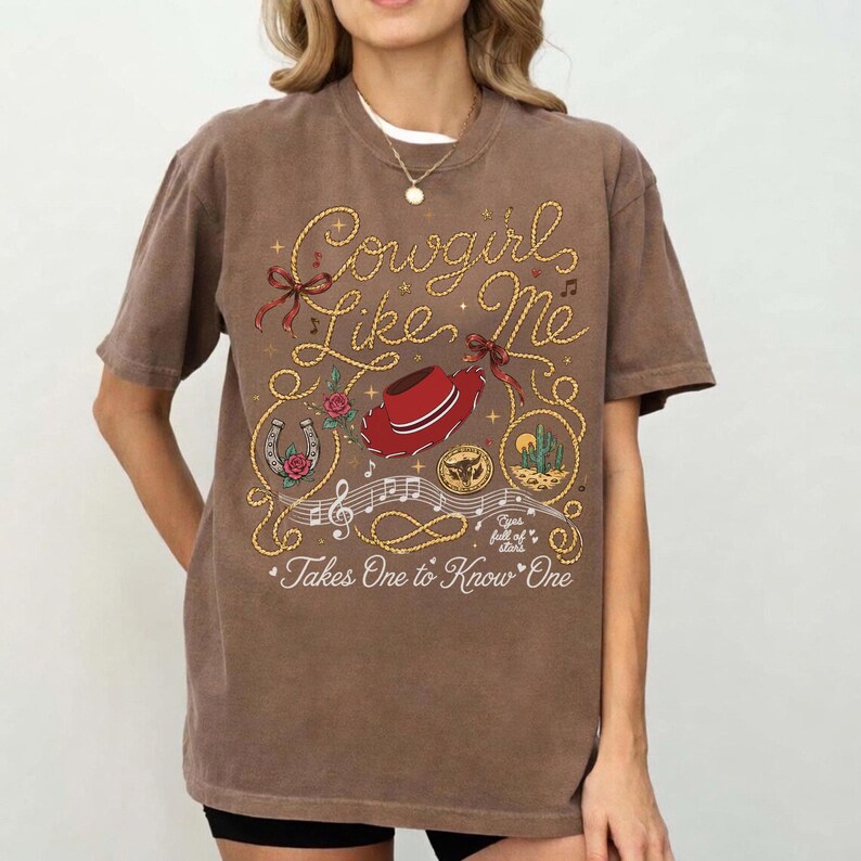 May include: A brown short-sleeve t-shirt featuring a graphic print with the text Cowgirl Like Me and Takes One to Know One. The design includes a red cowboy hat, a horseshoe with a rose, a cactus, a gold coin, and musical notes in gold rope.