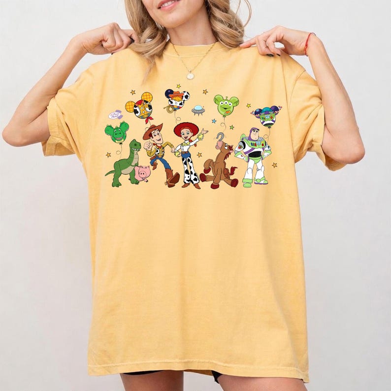 May include: Yellow t-shirt featuring a colorful graphic of Toy Story characters, including Woody, Buzz Lightyear, and Jessie. The design includes balloon-shaped versions of the characters and stars. The shirt is a comfortable, casual style.