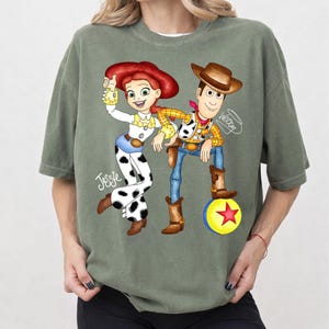 May include: Sage green t-shirt with a cartoon illustration of Jessie and Woody from Toy Story. Jessie is in a white and black cow-print outfit, and Woody is in a cowboy outfit. A yellow ball with a red star is also in the image.
