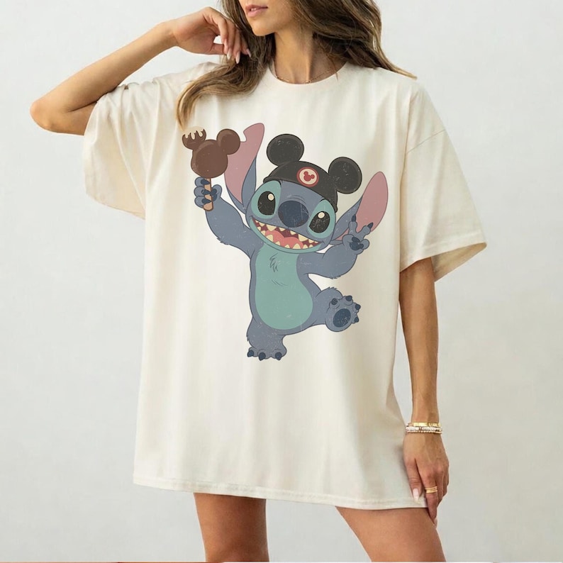 May include: Cream-colored oversized t-shirt featuring a cartoon illustration of Stitch from Lilo & Stitch. Stitch is depicted in blue and teal, wearing a Mickey Mouse hat and holding a Mickey Mouse-shaped ice cream. The design is centered on the shirt.