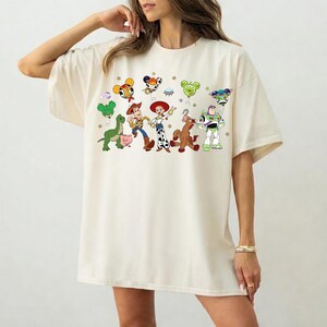 May include: Cream-colored oversized t-shirt featuring a colorful cartoon graphic of characters from the Toy Story movie. The graphic includes Woody, Buzz Lightyear, and other characters. The shirt has short sleeves and a relaxed fit.