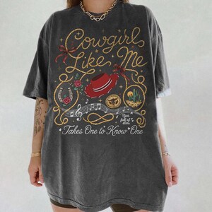May include: A charcoal gray short-sleeve t-shirt featuring a graphic print with the text Cowgirls Like Me and Takes One to Know One. The design includes a red cowboy hat, a horseshoe, a cactus, musical notes, and rope illustrations.