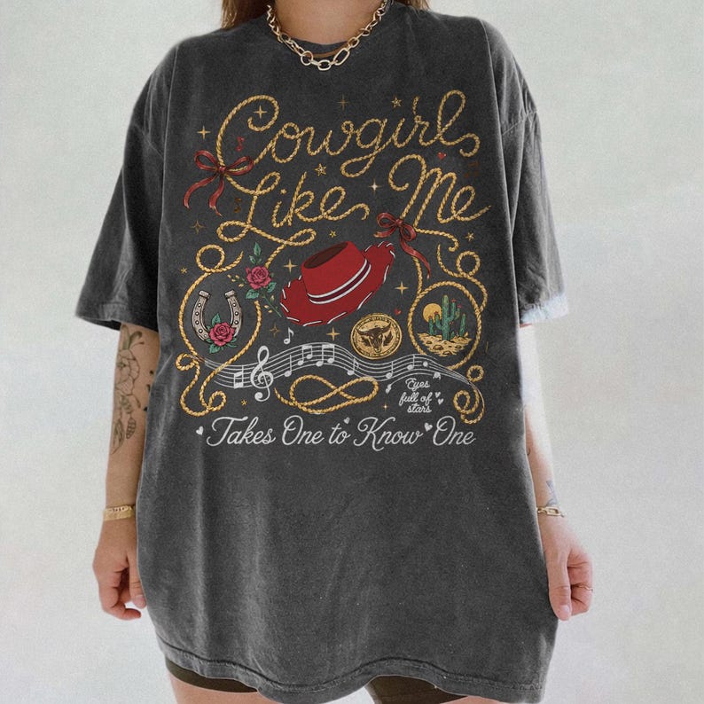 May include: A charcoal gray short-sleeve t-shirt featuring a graphic print with the text Cowgirls Like Me and Takes One to Know One. The design includes a red cowboy hat, a horseshoe, a cactus, musical notes, and rope illustrations.